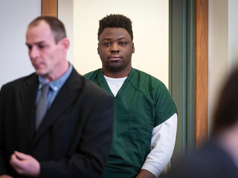 Two individuals in a room, one wearing a green jumpsuit standing in the background, and another in a suit in the foreground.