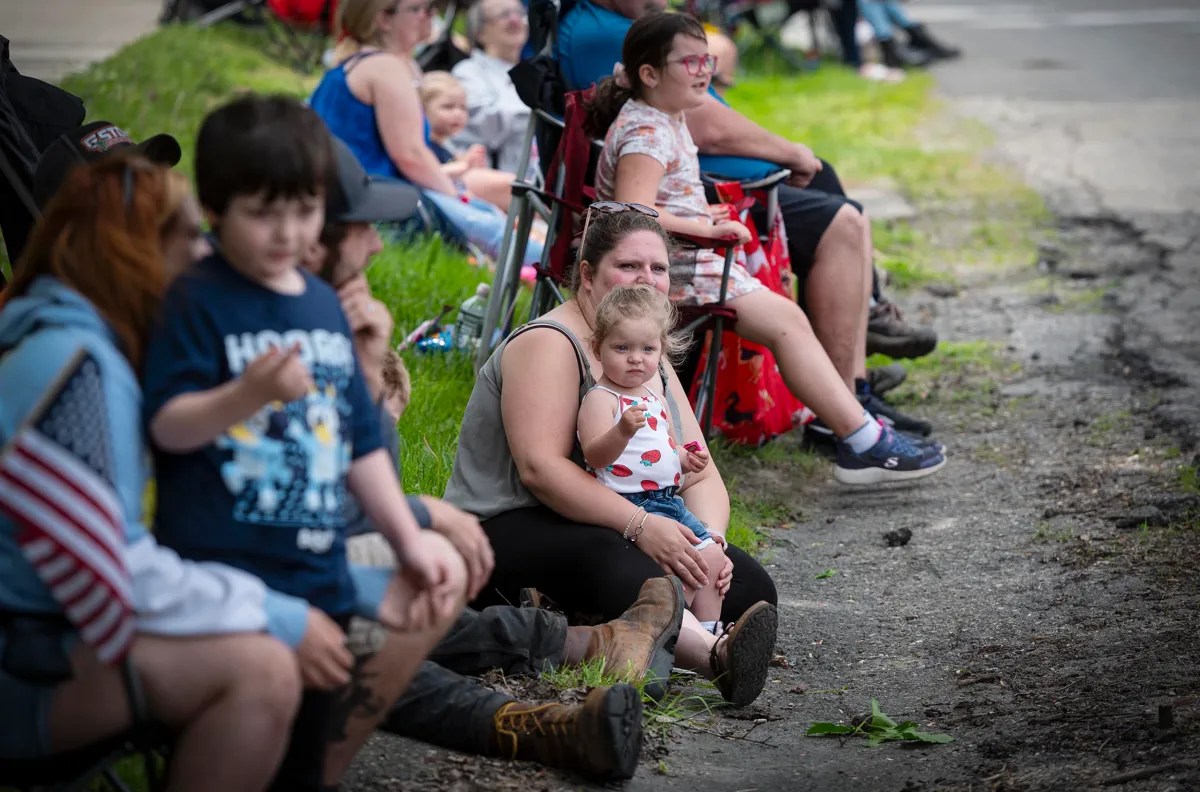 People seated outdoors on a sidewalk, with some sitting on portable chairs and others on the ground. An adult is holding a small child.