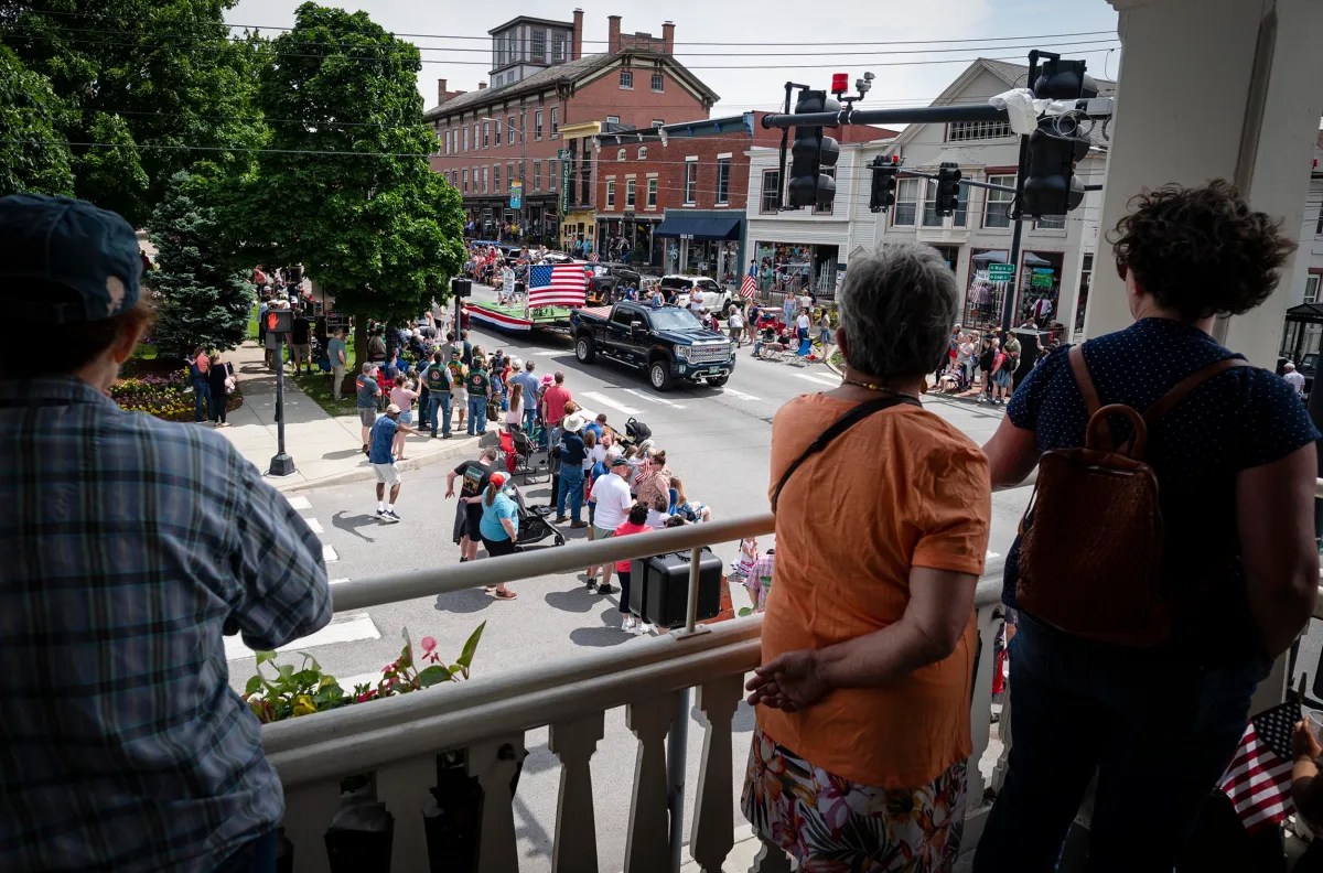 A crowd gathers along a street during a daytime parade, with people watching from a porch. A truck with an American flag leads the procession, and various buildings and shops are visible in the background.