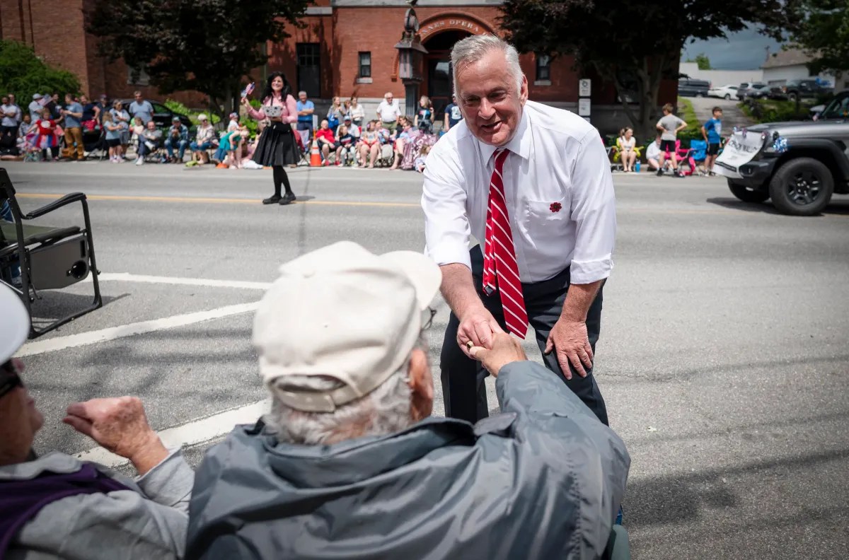 A man in a white shirt and red striped tie shakes hands with an elderly person on the street during an outdoor event. Bystanders and a brick building are visible in the background.