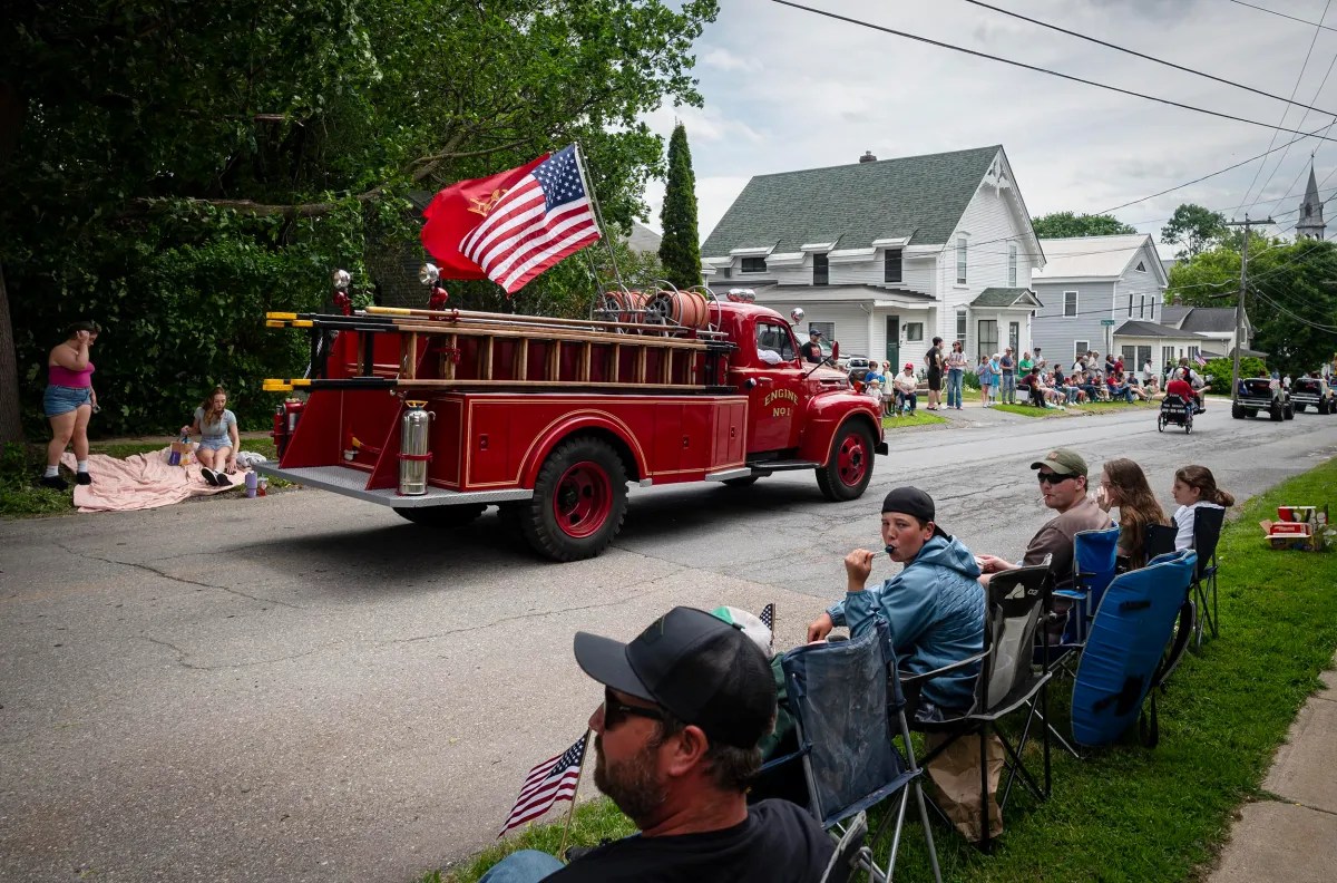 A vintage red fire truck displaying American flags drives down a residential street in a parade. Spectators sit and stand on the sidewalk watching the scene.