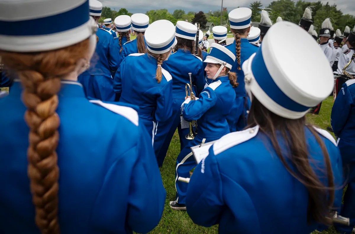 A marching band in blue uniforms and white hats is gathered outdoors. One band member, holding a brass instrument, looks back over their shoulder while others face away.