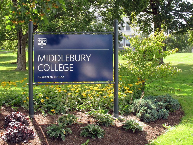 A blue sign reading "Middlebury College, Chartered in 1800" stands in a landscaped area with flowers and greenery, with trees and a building in the background.