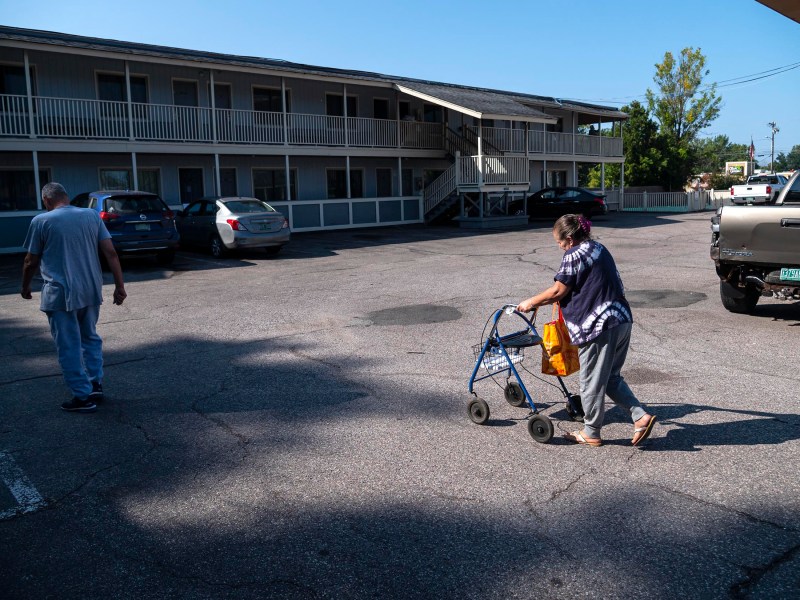 An elderly woman uses a walker in a parking lot of a two-story motel, while a man walks ahead of her.