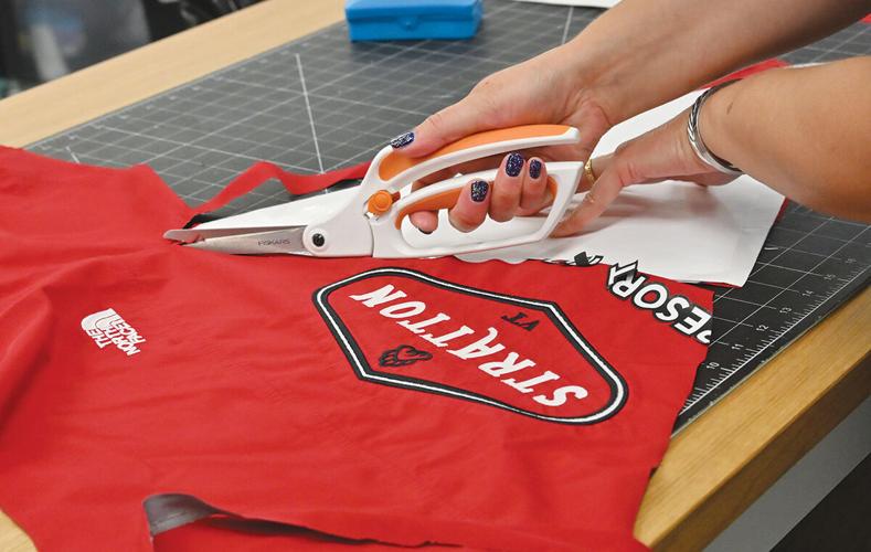A person cuts a red t-shirt with "STIRRATTON" and a logo using white fabric scissors on a black grid cutting mat.