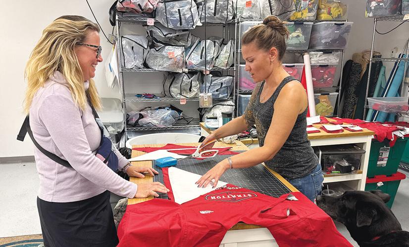 Two women work together at a table, cutting and assembling fabric pieces in a workshop filled with shelves of bags and materials.