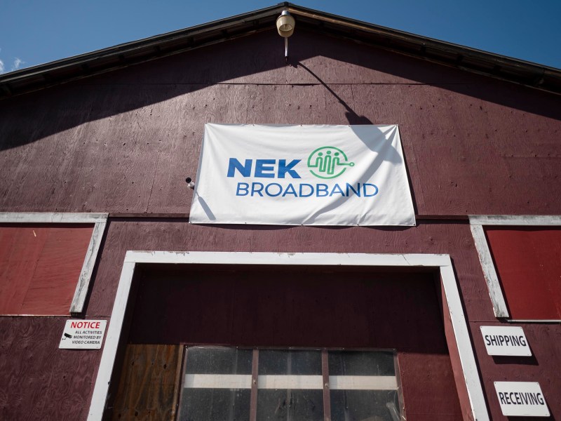 A large sign reading "NEK Broadband" is displayed above the entrance of a brown warehouse with shipping and receiving signs by the door.
