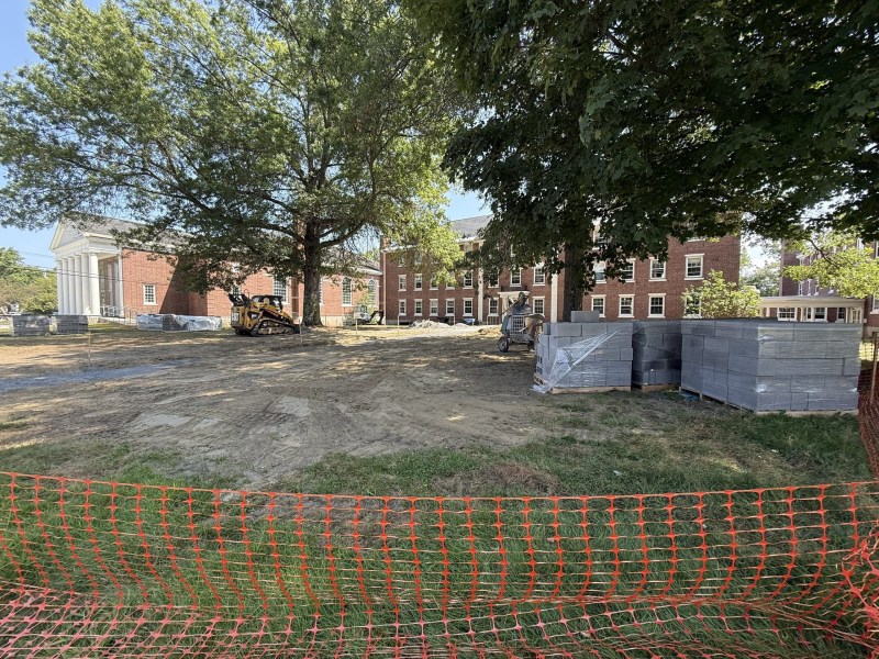 A construction site with heavy machinery, stacks of building materials, and an orange safety fence in front of brick buildings and trees.