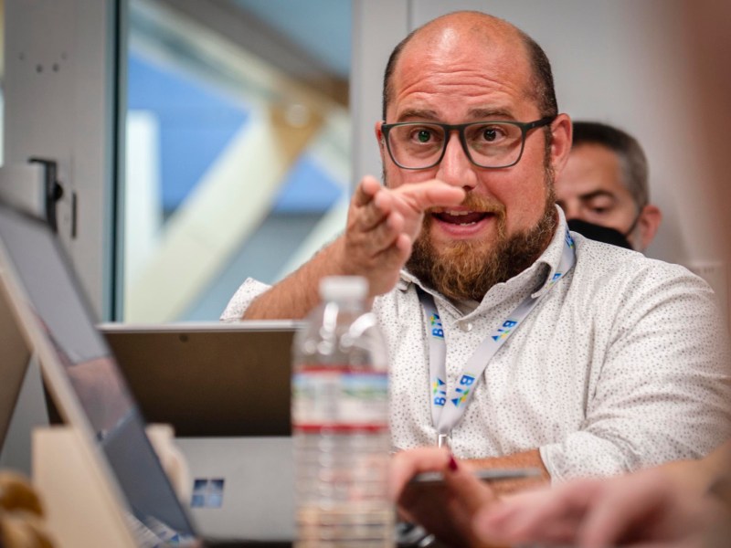 A man with glasses and a beard gestures while speaking during a meeting, seated at a table with laptops and a water bottle.
