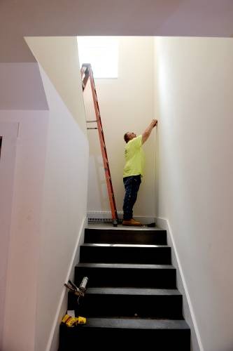 A person stands on a ladder positioned at the top of a staircase, working on a wall near a window with tools placed on the stairs.