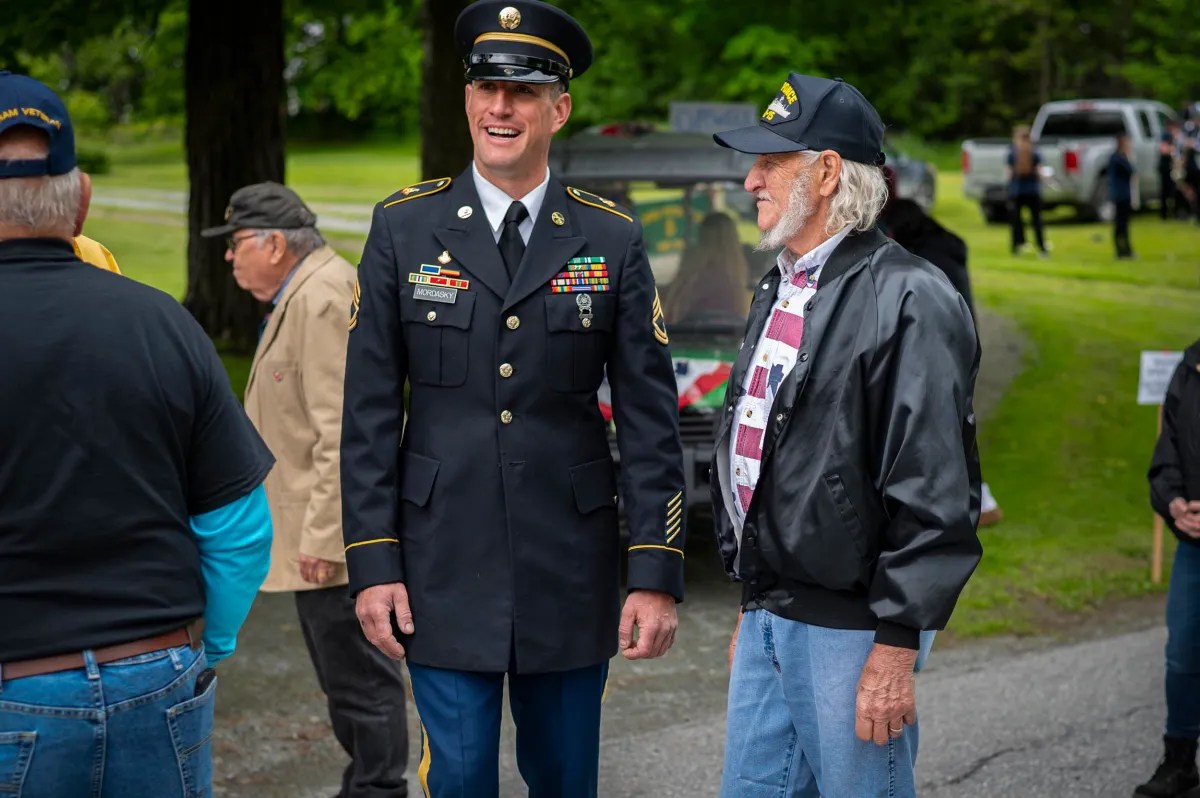 A man in a military dress uniform stands and smiles while conversing with another man in casual clothing at an outdoor gathering.
