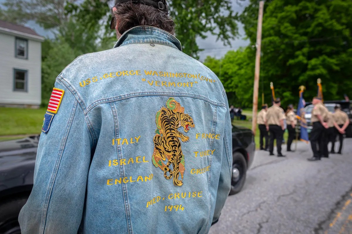 A person wearing a denim jacket with embroidery of a tiger and military-related text stands on a street near a group of people in uniform.