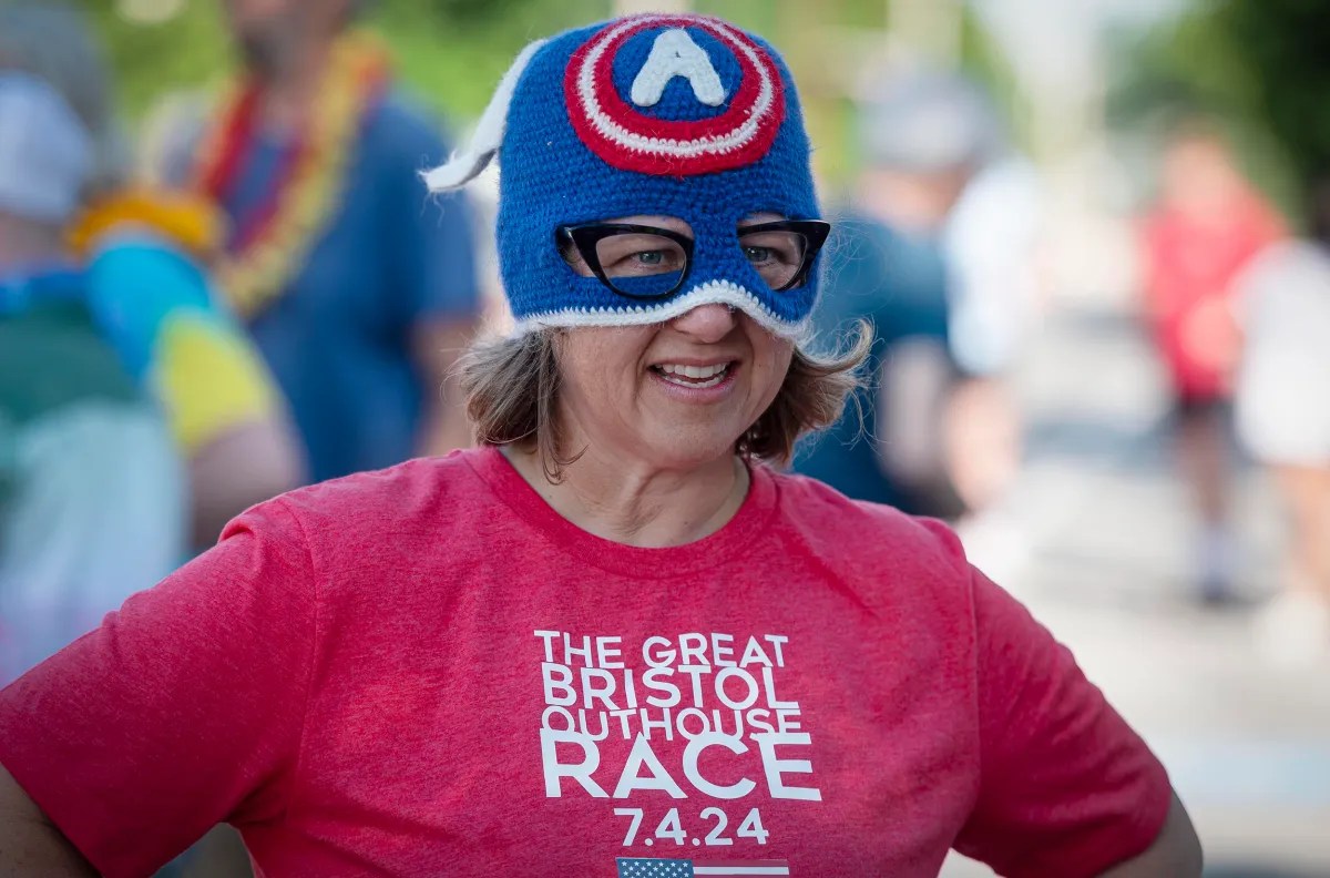 A person wearing a red "The Great Bristol Outhouse Race 7.4.24" shirt and a blue and red knit hat with a white "A" emblem, smiling outdoors.