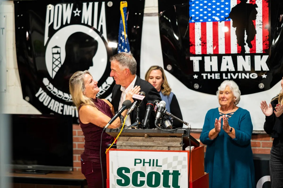 A man is speaking at a podium with several microphones. Banners behind him say "POW-MIA" and "Thank the Living." A woman next to him holds his arm. People in the background are clapping.