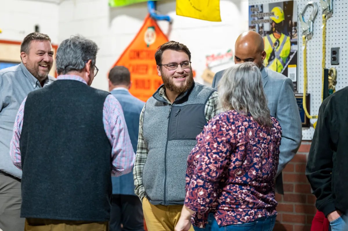 A group of people are having a conversation in an indoor setting with safety equipment and signage in the background.