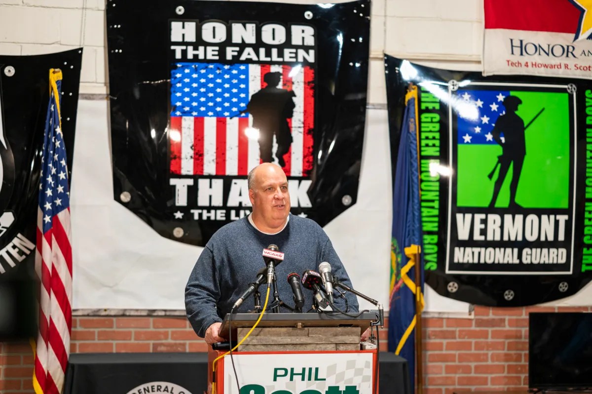 A man speaks at a podium with multiple microphones, standing before banners honoring military service. An American flag is visible to his right.