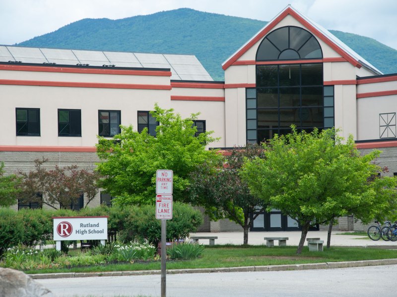 A beige and rust-colored building with large windows surrounded by trees and a lawn.