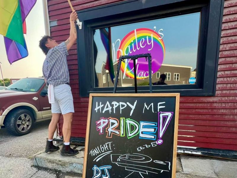 A person raises a pride flag outside Bailey’s Place, with a colorful “Happy MF Pride!” sign and a DJ announcement displayed on a chalkboard.
