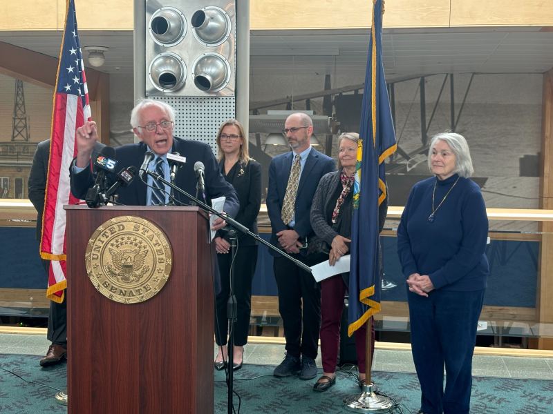 Senator speaks at podium with U.S. Senate seal, flanked by American flags, while five people stand behind him and one woman stands to the side.