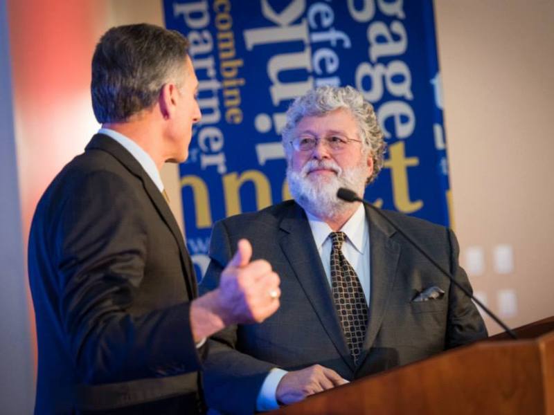 Two men in suits stand at a podium having a conversation, with one facing forward and the other giving a thumbs up. Blue and yellow signage is visible in the background.