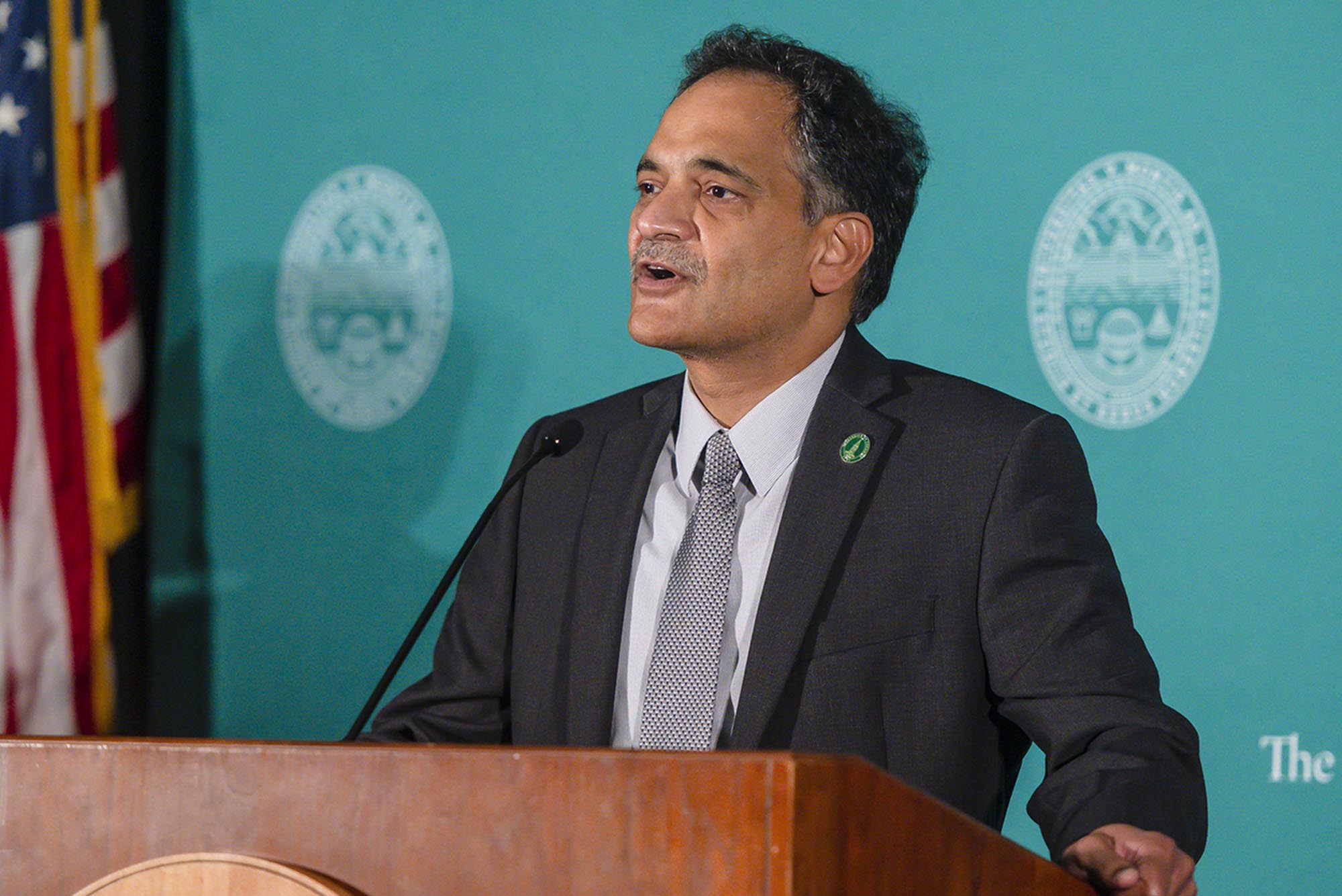 A man in a suit stands at a podium with a microphone, speaking in front of a teal backdrop featuring a circular emblem and an American flag.