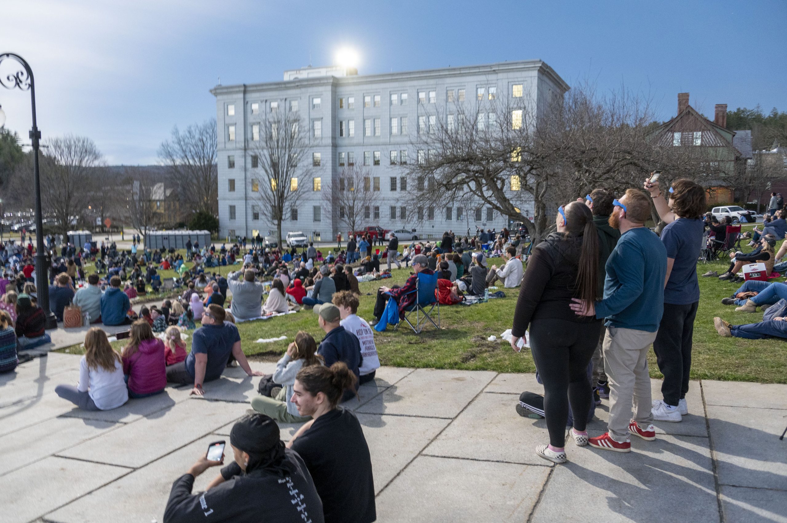 Crowd of people gathered on a grassy field in front of a building, with some standing and others seated on the ground.
