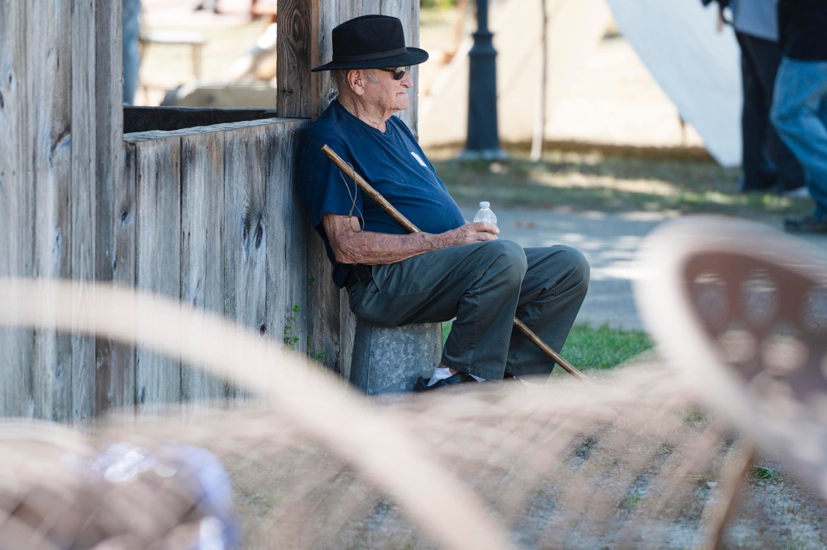 An elderly man wearing a black hat and sunglasses sits on a stone by a wooden wall, holding a water bottle and a cane.