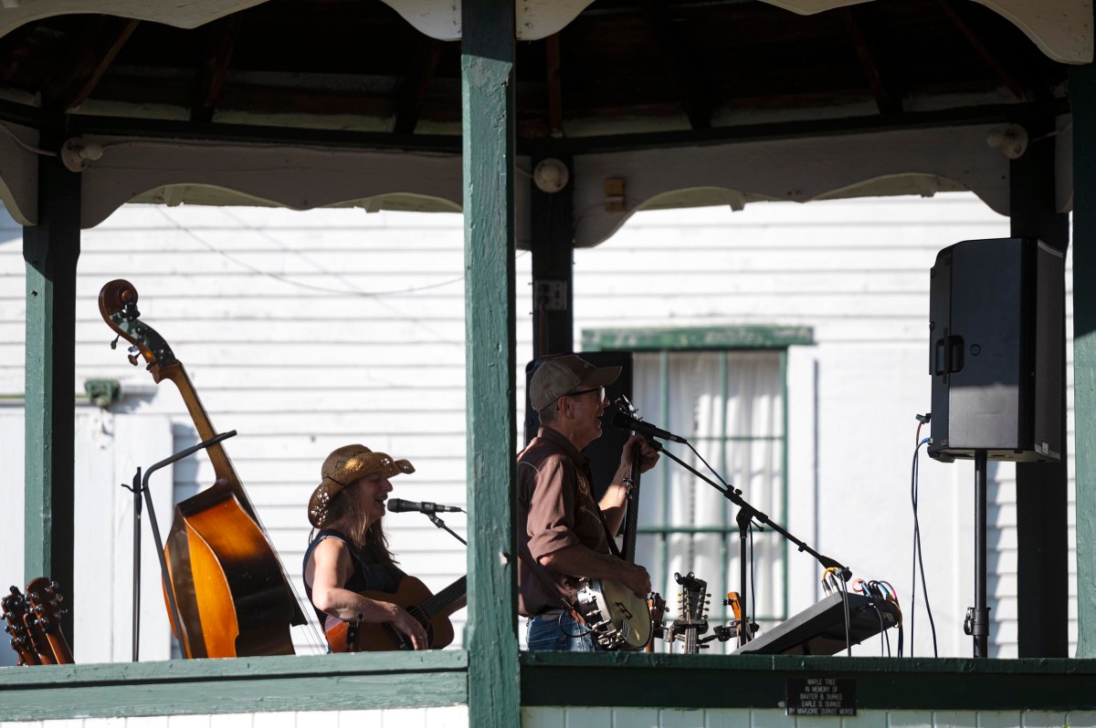 Two musicians play instruments and sing into microphones under a shaded gazebo, with a white wooden building in the background.