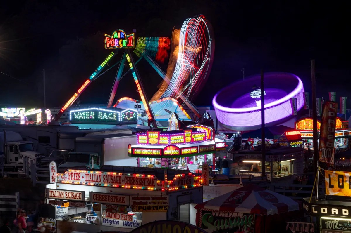 Nighttime scene at a fair with brightly lit food stalls and colorful amusement rides in motion, including a spinning ride and a Ferris wheel.