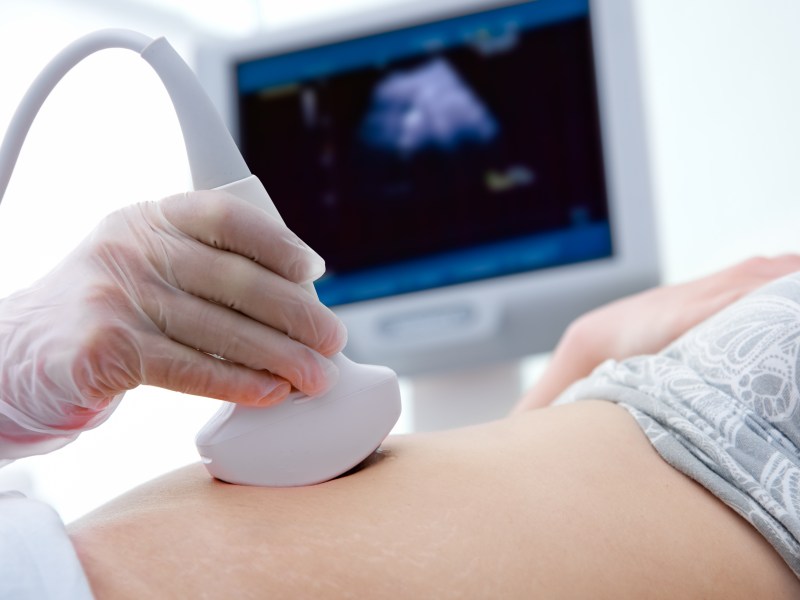 A doctor uses an ultrasound machine on a patient's stomach with a screen in the background.