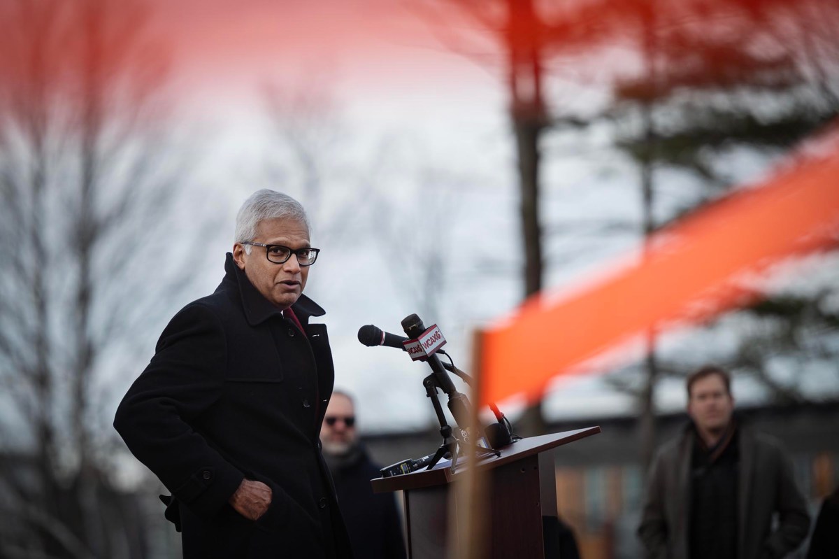 A person in a black coat speaks at a podium with microphones outdoors on a cloudy day. An orange ribbon is visible in the foreground.