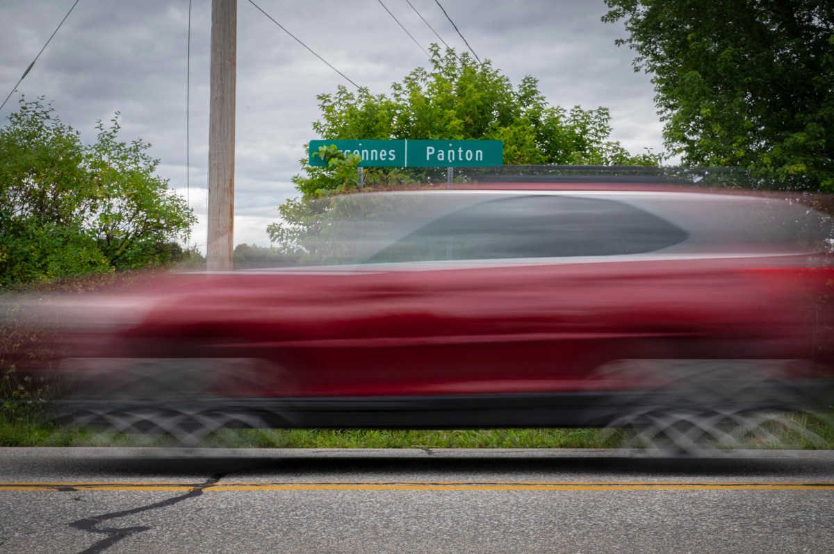 A red SUV speeds past a green road sign reading "Paxton" and "Nasonnes" on an overcast day, with trees and power lines in the background.