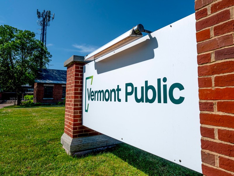 A white sign reading "Vermont Public" is mounted between two brick pillars outside, with a grassy area and a communication tower in the background.