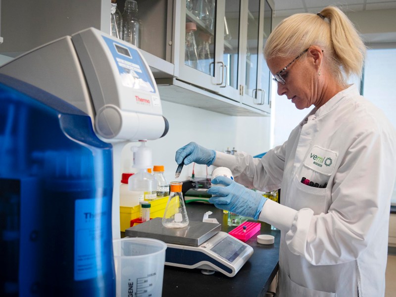 A scientist in a lab coat and gloves measures liquid into a beaker on a scale in a laboratory setting with scientific equipment and supplies.