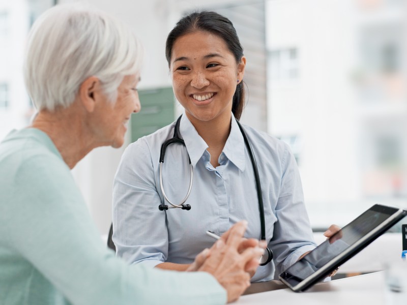 A healthcare professional with a stethoscope shows a tablet to an older woman during a consultation in a bright office.