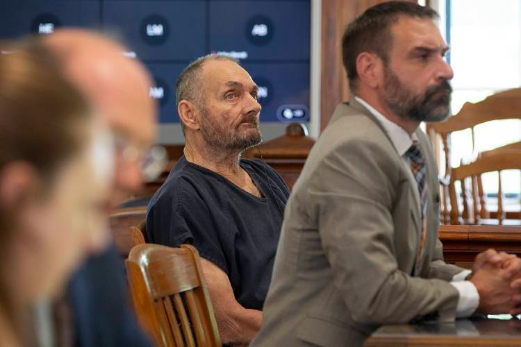 A man in a dark shirt sits beside a suited attorney in a courtroom, with other people and empty chairs visible in the background.