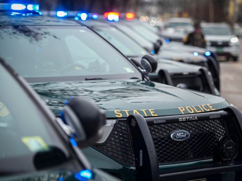 A row of state police vehicles with flashing lights line a street in an urban area.