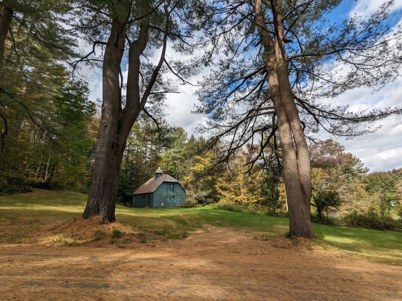 A small green cabin sits on a grassy clearing among tall trees under a partly cloudy sky.