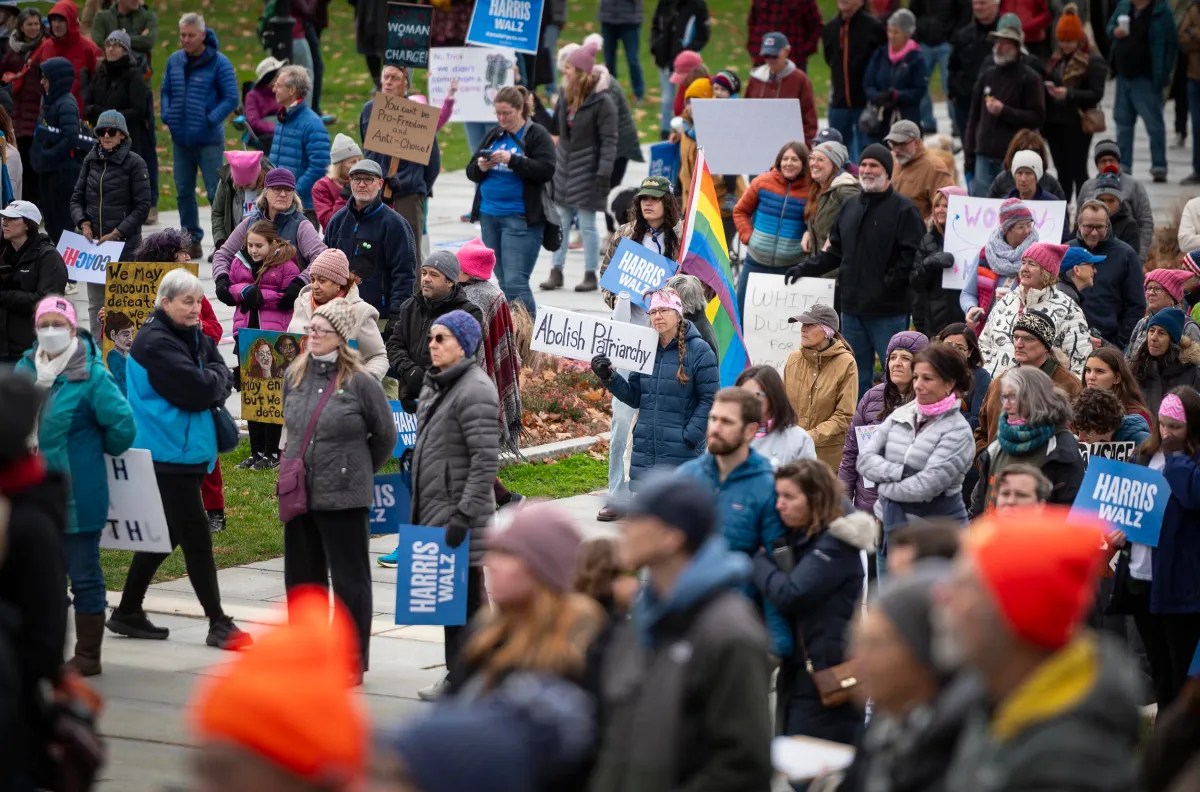 A large group of people holding signs at an outdoor protest. Various slogans are visible, including "Abolish Patriarchy." Some attendees wear pink hats and winter clothing.