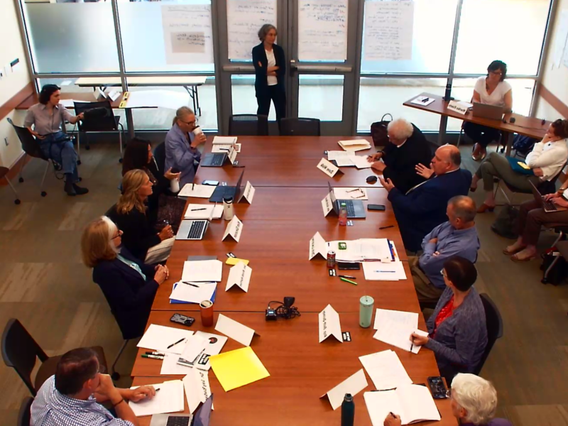 A group of people sits around a conference table in a meeting room, with one person standing and presenting in front of large windows and flip charts.